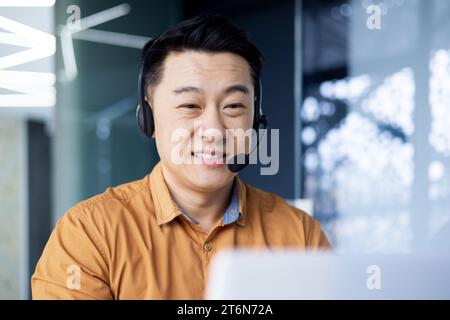 Uomo coreano di buon umore lavoratore d'ufficio in cuffia siede alla scrivania di fronte a un laptop in un ufficio moderno, un uomo asiatico lavora in un call center consiglia i clienti, tocca una tastiera. Foto Stock