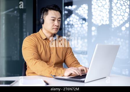 Impiegato coreano concentrato in ufficio con cuffie sedute alla scrivania di fronte al laptop in un ufficio moderno, uomo asiatico che lavora in un call center che consiglia i clienti, toccando la tastiera. Foto Stock