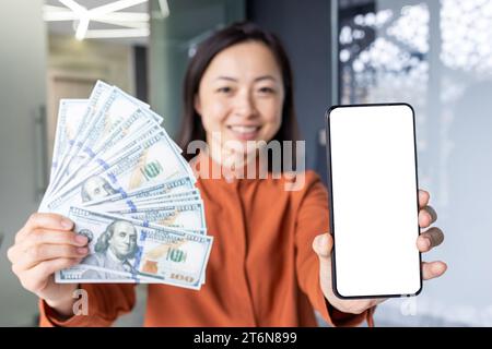 Eccitata giovane donna cinese che ha un grande smartphone con una cella bianca a schermo vuoto per i mockup e banconote in denaro contante in mano, seduto e in ufficio. Cashback, concetto di trasferimento di denaro online semplice. Foto Stock