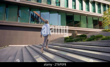 Vista sul retro donna d'affari di mezza età manager CEO leader imprenditore avvocato professionista donna irriconoscibile che cammina all'aperto verso l'edificio degli uffici Foto Stock