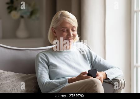 Donna matura sorridente che tiene il telefono, seduta su un comodo divano Foto Stock