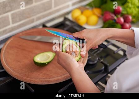 Primo piano le mani di una donna che tiene in mano un frutto di avocado e lo sbuccia, in piedi al bancone della cucina, preparando un'insalata sana per cena. Uno stile di vita sano Foto Stock