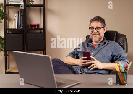 Uomo di mezza età sorridente davanti allo smartphone, che lavora comodamente da un ufficio domestico ben organizzato. Foto Stock