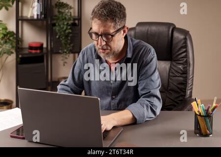 Uomo concentrato che lavora diligentemente sul suo laptop in un ambiente di ufficio domestico, esemplificando la professionalità del lavoro remoto. Foto Stock