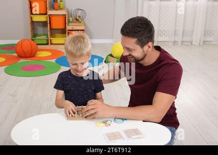 Sviluppo di competenze motorie. Padre felice che aiuta suo figlio a giocare con geoboard e fasce di gomma al tavolo bianco in camera Foto Stock