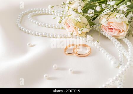 Organizzazione delicata del matrimonio con due anelli nuziali in oro su sfondo raso crema con perle e fiori. cartolina. invito. copertura Foto Stock