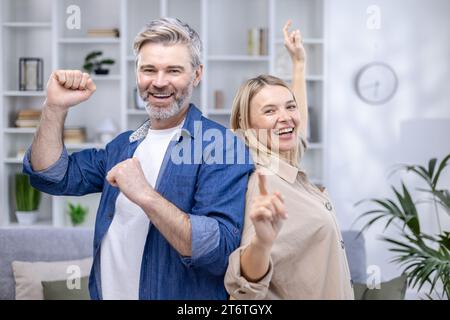 Coppia anziana felice che balla nel loro accogliente soggiorno, sorridente guardando la macchina fotografica, felice matrimonio lungo, marito e moglie trascorrono il fine settimana a casa. Foto Stock