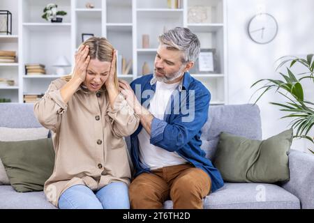 Coppia anziana seduta sul divano in salotto, moglie che si strofina la testa con le mani, soffre di grave emicrania, marito che la abbraccia, la conforta, offre aiuto. Foto Stock