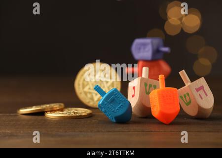 Dreidels con lettere e monete ebraiche su un tavolo di legno contro luci festive sfocate, concentrazione selettiva. Spazio per il testo. Tradizionale gioco Hanukkah Foto Stock