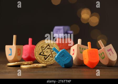 Dreidels con lettere e monete ebraiche su un tavolo di legno contro luci festive sfocate, concentrazione selettiva. Tradizionale gioco Hanukkah Foto Stock