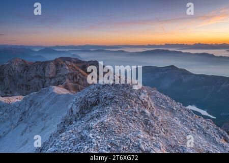 Prima luce del sole che colpisce i pendii rocciosi delle montagne con il lago Tovel e i fondali nebbiosi della valle. Alta alba alpina nelle Dolomiti di Brenta. Foto Stock