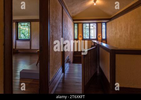 La Frederick C. Robie House conduce, attraverso un pianerottolo con soffitto inclinato e una fila di finestre a vetri piombati a motivi geometrici a Chicago, Illinois, Stati Uniti, alle stanze private all'ultimo piano. Frank Lloyd Wright ha dotato interamente di finestre a vetri piombati il livello del sottotetto, noto come belvedere, della casa in stile Prairie, costruita nel 1909/1910. Una ringhiera in legno tinteggiato scuro delimita la scala in basso in primo piano. Dal 2019, la casa è una degli otto edifici di Wright inseriti nella lista del Patrimonio mondiale dell'UNESCO. Foto Stock
