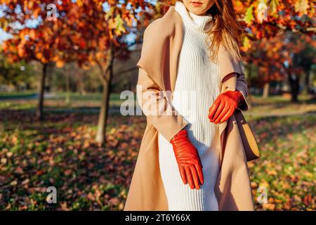 Guanti femminili in pelle arancione. Donna alla moda che indossa un elegante cappotto beige, abito a maglia bianca che cammina nel parco autunnale con borsa Foto Stock
