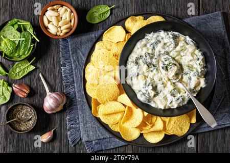 salsa cremosa di mandorle e spinaci di carciofi con patatine fritte in ciotola su piatto nero su tavola di legno scuro con ingredienti, vista orizzontale, posa piatta Foto Stock