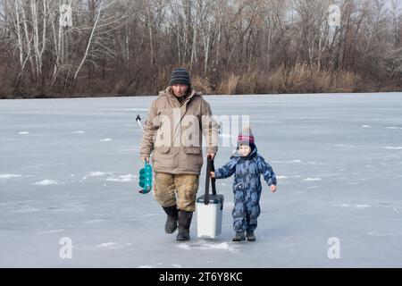 Nonno e nipote camminano lungo il fiume ghiacciato per la pesca invernale. Un uomo anziano tiene in mano un'esercitazione sul ghiaccio e una scatola da pesca, Th Foto Stock