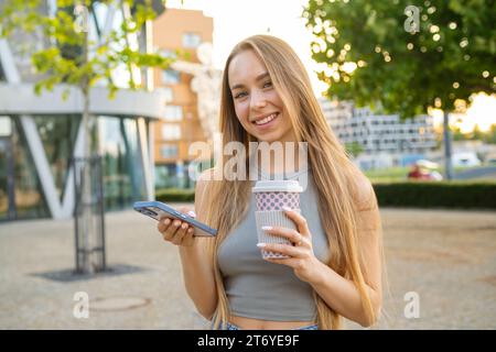 Giovane donna sorridente dai capelli biondi che controlla le app del telefono cellulare e beve caffè in strada Foto Stock
