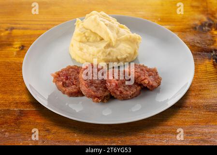 Fette di salsiccia cotta cotechino con purè di patate e foglie di salvia sul piatto su un tavolo di legno. Taglio della piastra con percorso di ritaglio incluso Foto Stock
