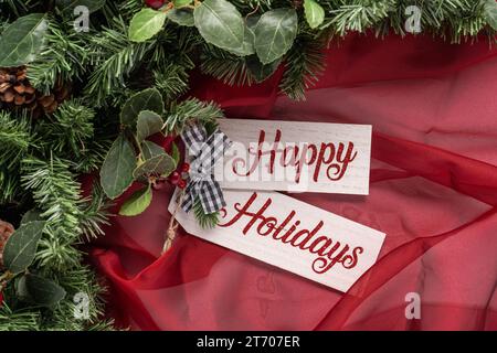 Testo Happy Holidays su tavola di legno con tessuto rosso trasparente, vegetazione e un piccolo arco a Vichy bianco e nero Foto Stock