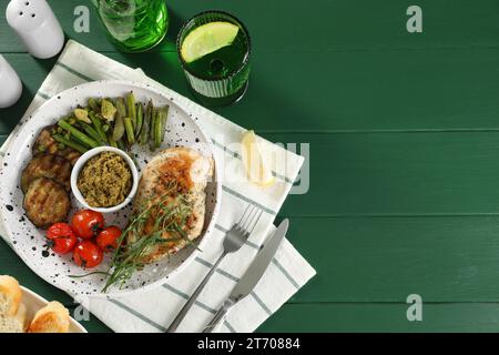 Gustoso pollo, verdure, bevanda con dragoncello e salsa al pesto servita su un tavolo di legno verde, piatti. Spazio per il testo Foto Stock