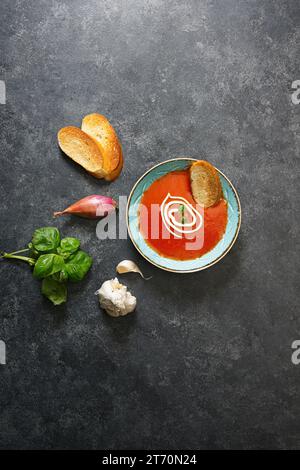 Crema di pomodoro e zuppa di basilico in un recipiente su sfondo nero con ingredienti intorno Foto Stock