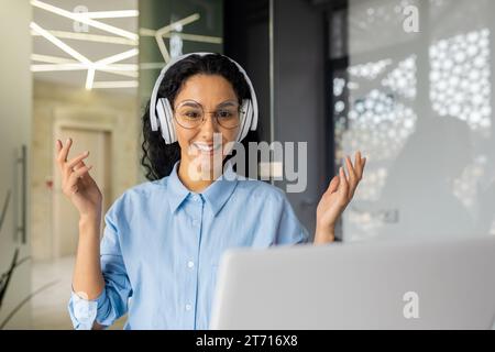 Donna indiana di buon umore, lavoratrice in ufficio con le cuffie, seduta alla scrivania di fronte al computer portatile in un ufficio moderno, donna ispanica che parla in videochiamata, sorride, gesti con le mani Foto Stock