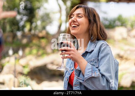 Una donna con una giacca in denim ride mentre si gusta un bicchiere di vino rosso, catturando un momento di pura gioia. Foto Stock