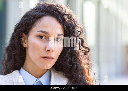 Ritratto ravvicinato di una bella lavoratrice latinoamericana in piedi fuori dall'ufficio alla luce del sole, incredibile studentessa bruna che guarda seriamente la fotocamera. Foto Stock