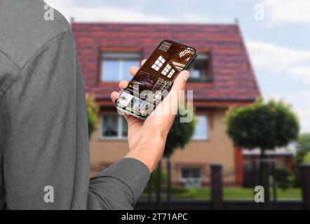 Uomo che utilizza un sistema di sicurezza domestica intelligente su un telefono cellulare vicino a casa all'aperto, primo piano. Dispositivo che mostra stanze diverse attraverso le fotocamere Foto Stock