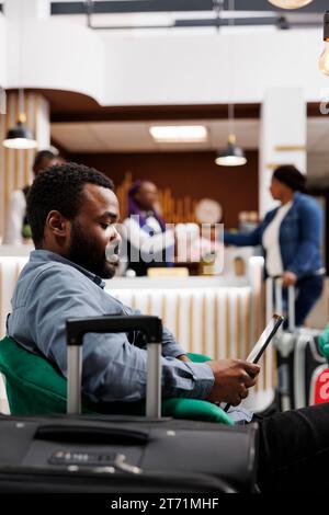 Viaggiatore afroamericano che utilizza un tablet digitale per il check-in in hotel, seduto con i bagagli nella lobby, tecnologia di check-in self-service nell'ospitalità. Turista di tipo nero in attesa del processo di registrazione Foto Stock