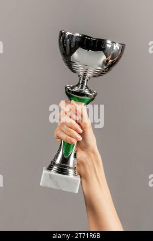 Anonima giovane donna sottile con dita sottili e curate che tengono in mano un trofeo lucente e che si manifesta su sfondo grigio Foto Stock