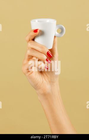 Anonimo giovane mano femminile sottile con unghie lucide e sottili che tengono in alto la tazza di caffè di luce su sfondo marrone Foto Stock