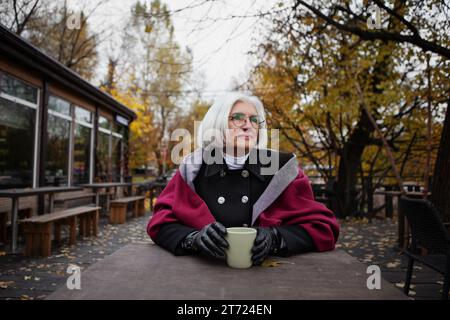 Elegante donna anziana con capelli grigi seduta al tavolo del caffè all'aperto nel parco autunnale con una tazza di tè o caffè, indossando abiti e occhiali da vista in modo confortevole Foto Stock