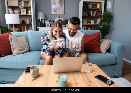 Sorpreso coppia felice che annuncia la gravidanza a parenti e amici durante una videochiamata. Giovani futuri genitori che utilizzano un computer portatile per condividere le loro Foto Stock