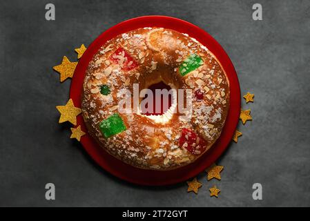 Vista dall'alto di Roscon de reyes su una targa rossa con stelle dorate. Torta spagnola "Three King" su sfondo grigio. Il concetto di "Kings Day" Foto Stock