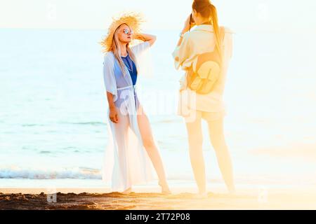 Il fotografo fotografa una bella bionda con un cappello di paglia la sera d'estate in riva al mare. Foto Stock