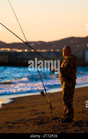 L'uomo anziano pesca con la canna da pesca sulla riva dell'oceano al tramonto. Tempo libero del pensionato. Foto Stock