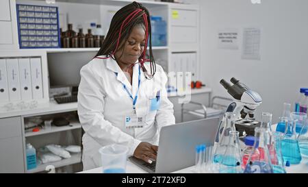 Scienziata afroamericana concentrata impegnata in una seria ricerca medica sul suo portatile in laboratorio, ritratto di un bellissimo adulto con trecce consegnare Foto Stock