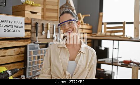 Bellissima giovane donna ispanica, un falegname sorridente, orgogliosa della sua officina di falegnameria, che indossa occhiali di sicurezza Foto Stock