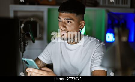 Bellissimo giovane latino, musicista tatuato, che esprime emotivamente la sua canzone sullo smartphone in mezzo alla notte buia nel suo studio musicale Foto Stock