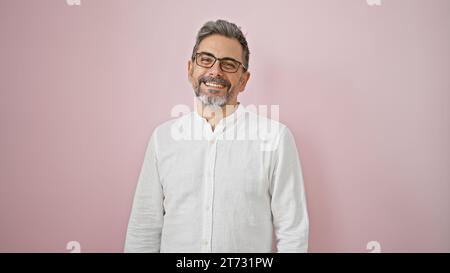 Uomo ispanico dai capelli grigi sicuro di sé che sorride con un sorriso affascinante, indossa occhiali, si erge su uno sfondo rosa isolato e irradia positività Foto Stock