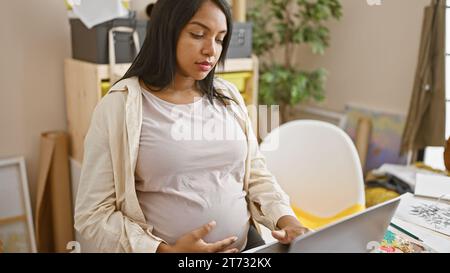 Giovane donna incinta, seduta al suo tavolo da studio d'arte, padroneggia la sua arte, dipingendo su tela con un portatile come guida. Foto Stock