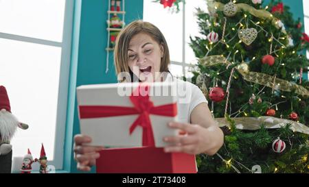 Donna ispanica di mezza età che apre il regalo con la faccia sorpresa a casa Foto Stock