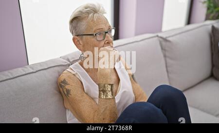 Donna anziana con i capelli grigi immersa nel profondo pensiero mentre si siede comodamente sull'accogliente divano di casa Foto Stock