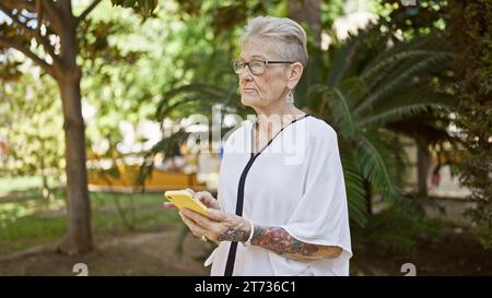 La donna anziana dai capelli grigi si concentra seriamente sul messaggio online, in mezzo al verde del parco illuminato dal sole, mostrando uno stile di vita maturo e esperto di tecnologia. Foto Stock