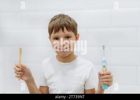 carino bambino di 8 anni che tiene in mano bambù e spazzolino elettrico per fare la scelta Foto Stock