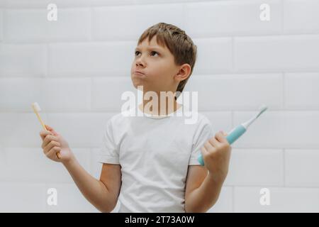 carino bambino di 8 anni che tiene in mano bambù e spazzolino elettrico per fare la scelta Foto Stock