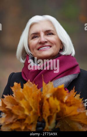 Primo piano Caucasica gioiosa vecchia dai capelli grigi con foglie autunnali sorridenti all'aperto. Felice maturo femmina Ritratto di anziana bella signora che si gode li Foto Stock