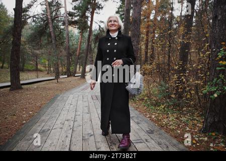 Donna anziana felice con i capelli argentati e un bel sorriso cammina attraverso il parco naturale nella foresta, rallegra e gode della bellezza della natura, genuina e sincera Foto Stock