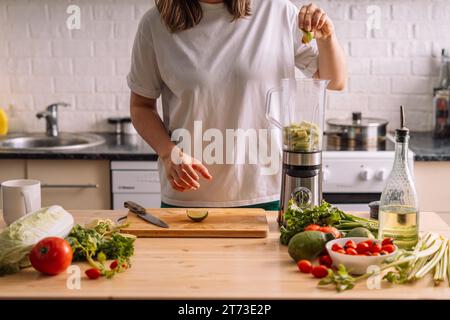 La signora in cucina spreme una calce in un frullatore su un tavolo di legno su cui ci sono verdure fresche Foto Stock