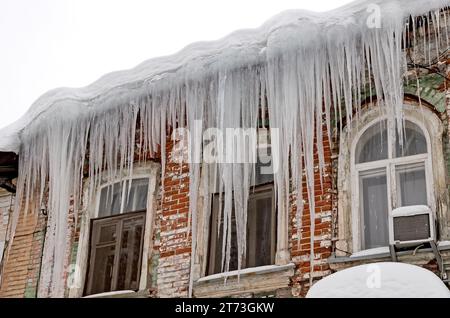 Enormi ghiaccioli affilati pendono pericolosamente dal tetto di una vecchia casa. Foto Stock